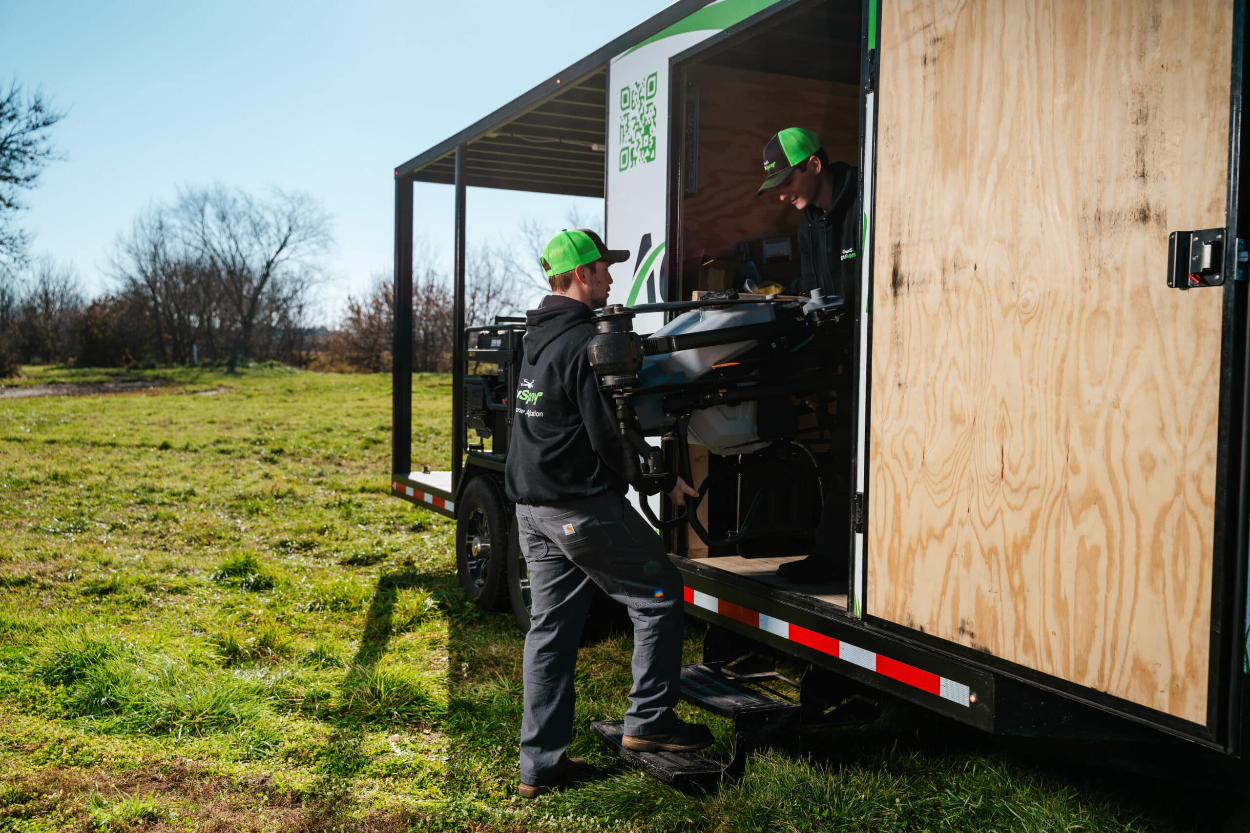 Two technicians in green caps and black jackets operating a drone from a mobile trailer workstation in a field.
