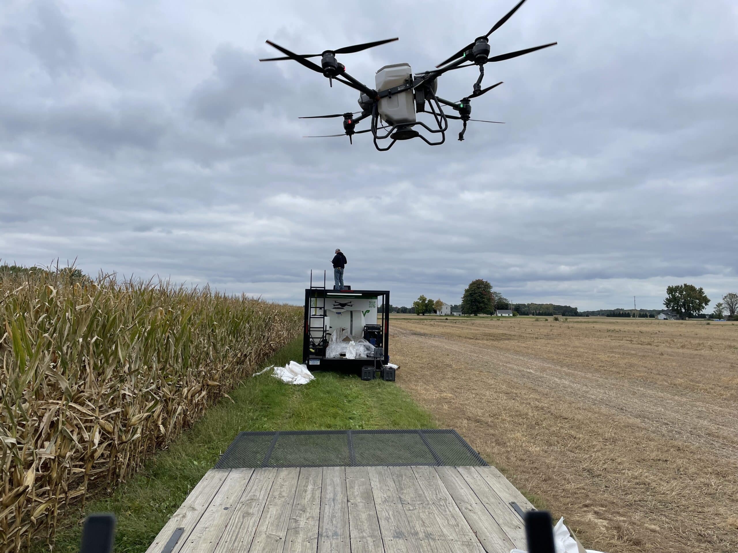 A commercial drone flies near an operator on a trailer by a cornfield, symbolizing modern technology in traditional farming.