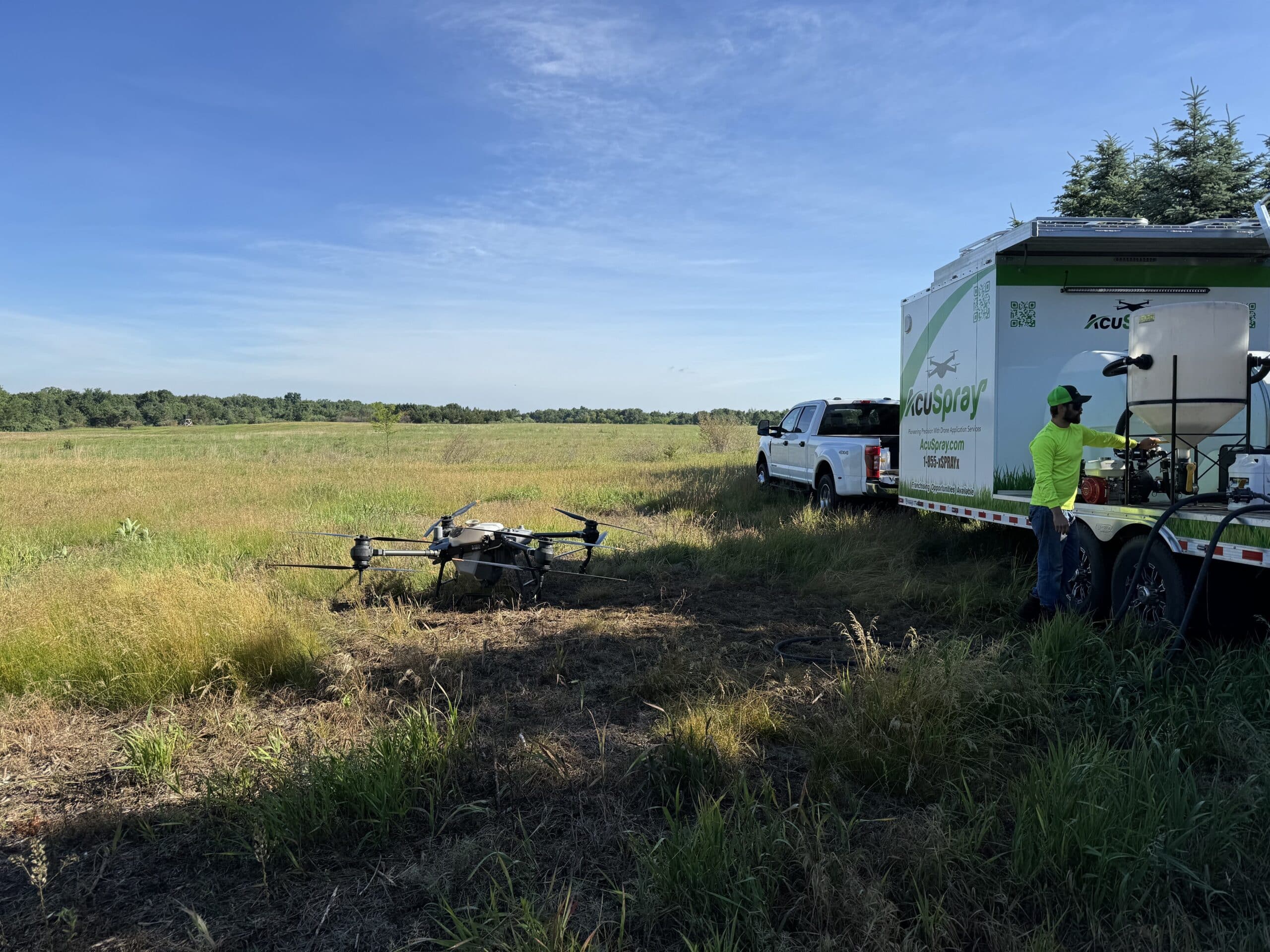 A technician prepares a drone next to an AcuSpray trailer in a field, illustrating the use of drone technology in precision agriculture for crop monitoring and treatment.